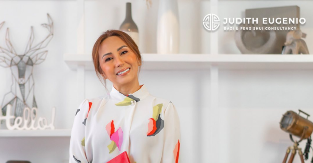 Smiling woman in a patterned blouse stands in front of white shelves adorned with decorative items. A logo at the top right reads "Judith Eugenio BaZi & Feng Shui Consultancy," where ancient Chinese metaphysics guide your personal energy reading and Ba Zi destiny forecast.