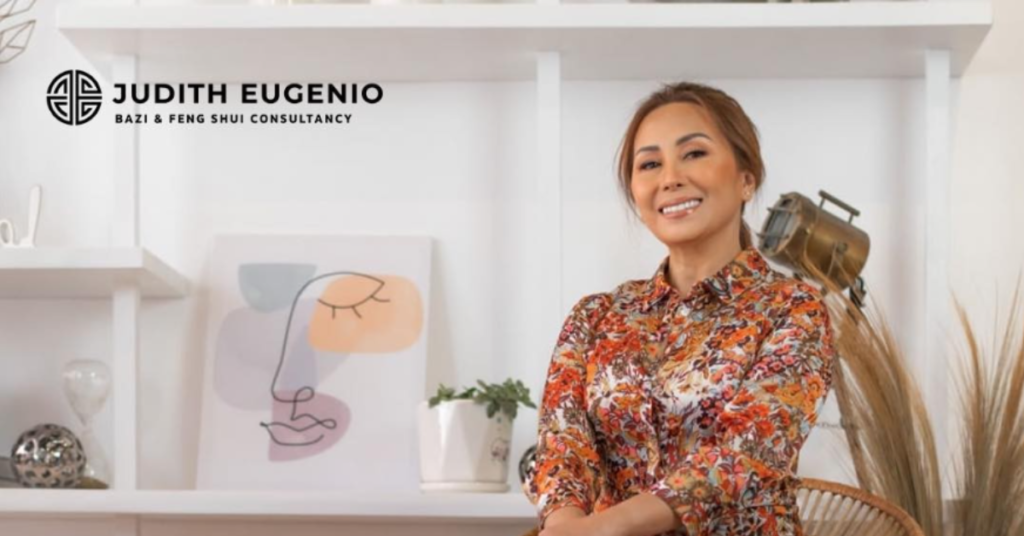 A woman in a floral shirt sits on a chair with a smile, framed by decor items and an abstract art piece. The text “Judith Eugenio Bazi & Feng Shui Consultancy” highlights her expertise in harnessing the Cosmic Trinity for prosperity and harmony.