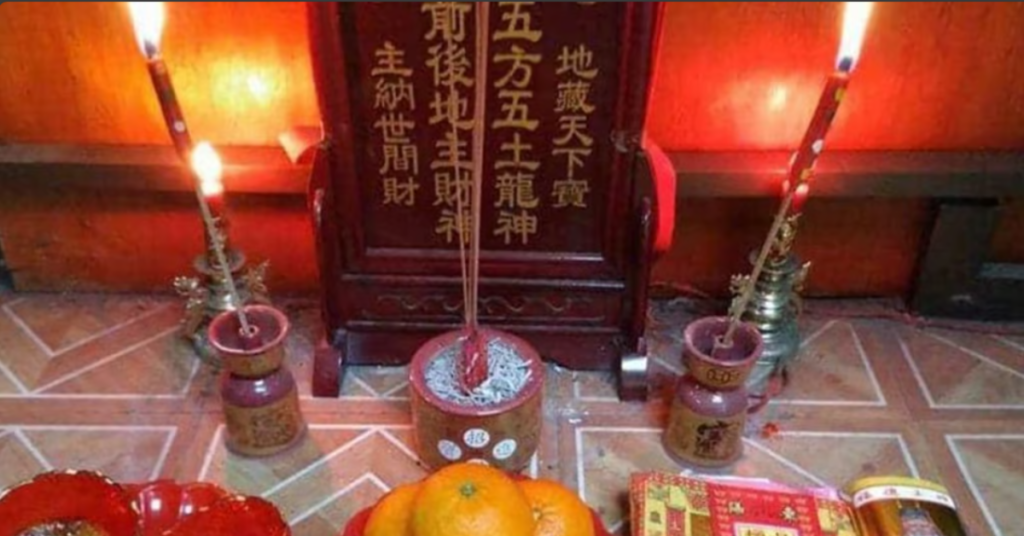 An altar with Chinese inscriptions, featuring three lit incense sticks, candles, oranges, and red packets on a tiled surface.