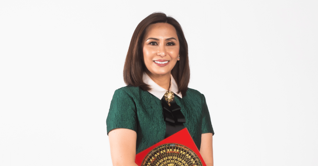 Person smiling while holding a decorative item with a circular pattern, wearing a green dress with a white collar against a plain white background.