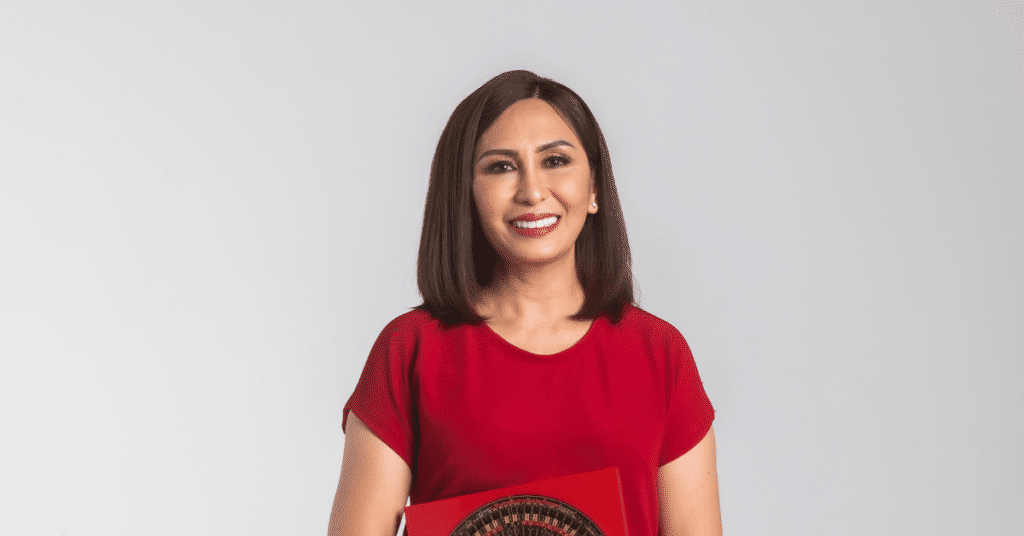 Person in a red shirt holding a red folder, smiling in front of a plain white background.