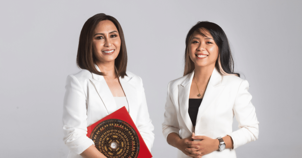 Two women in white blazers stand smiling against a gray background. One holds a red item with circular detailing.