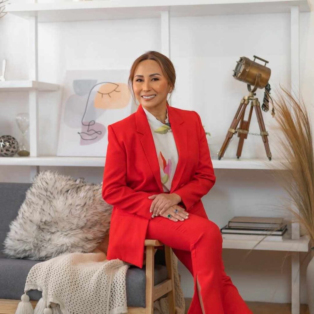 A woman in a bright red suit sits on a gray sofa in a modern, stylish room—a feng shui master offering spiritual guidance amid elegant shelves, decor items, and a tripod lamp in the background.
